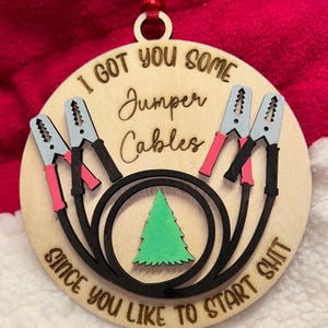 May include: A wooden Christmas ornament with the text "I GOT YOU SOME JUMPER CABLES SINCE YOU LIKE TO START SHIT." The ornament features a design of jumper cables, a green Christmas tree, and a red ribbon for hanging.