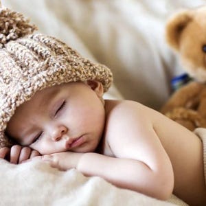 May include: A sleeping baby wearing a knitted brown hat with a pom-pom, resting on a soft, white blanket. A brown teddy bear is in the background. The baby's eyes are closed, and the skin is smooth.