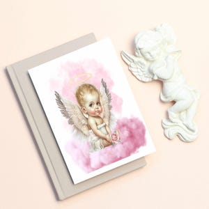 Puede incluir: Ilustración en acuarela de un querubín con alas, un halo y una rosa, sobre un fondo de nube rosa. Una figura de ángel blanco está al lado de la ilustración. La imagen está sobre un libro gris claro.