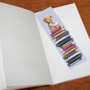 May include: A bookmark featuring a child reading atop a stack of books. The bookmark has a light purple background and is placed inside an open book. The books are various colors, including purple, yellow, and green.