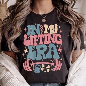 May include: Black t-shirt with the text "IN MY LIFTING ERA" in retro-style lettering. The design includes a barbell, kettlebell, and stars in pink, blue, yellow, and white. The shirt has a crew neck.