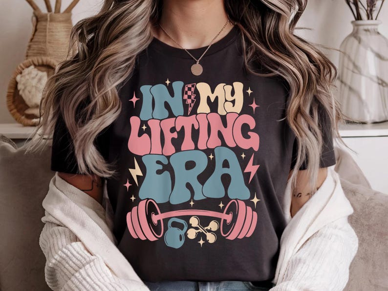 May include: Black t-shirt with the text "IN MY LIFTING ERA" in retro-style lettering. The design includes a barbell, kettlebell, and stars in pink, blue, yellow, and white. The shirt has a crew neck.