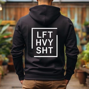 May include: Black hooded sweatshirt with the text "LFT HVY SHT" in a white square. The sweatshirt is a solid black color. The background is a blurred outdoor setting with plants.