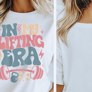 May include: White t-shirt with the text "IN MY LIFTING ERA" in retro-style lettering. The design includes a barbell, weights, and lightning bolts in pastel colors. The shirt is a casual wear item.