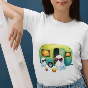 K&ouml;nnte beinhalten: Wei&szlig;es T-Shirt mit einer skurrilen Illustration eines gr&uuml;n-gelben Wohnwagens und zwei Geisterfiguren mit herzf&ouml;rmigen Sonnenbrillen. Bunte Strandb&auml;lle sind um die Geister und den Wohnwagen herum verstreut. Das Shirt ist aus weichem Material.