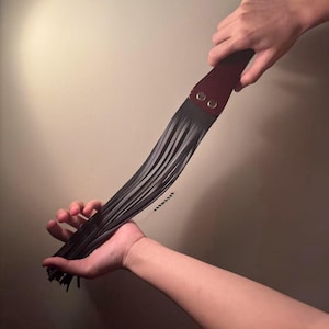 Pu&ograve; includere: Un frustino in pelle nera con un manico bordeaux fissato con due rivetti argentati. I fili del frustino sono tenuti in due mani su uno sfondo neutro.