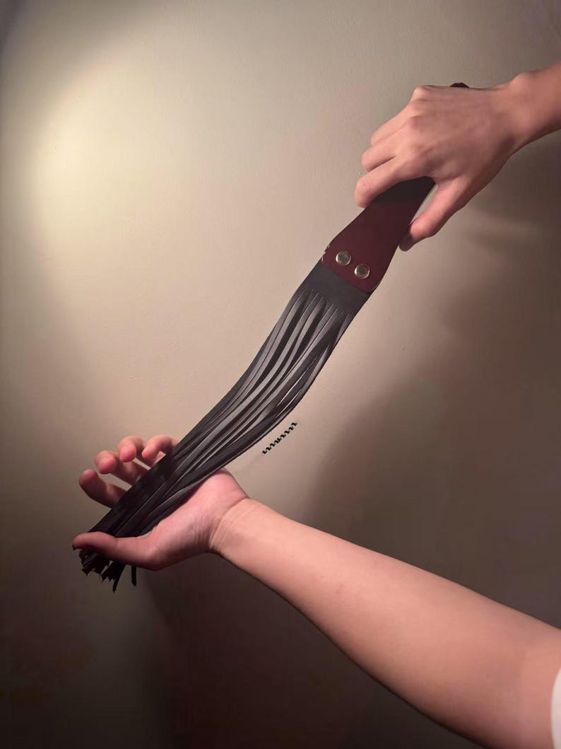 Pu&ograve; includere: Un frustino in pelle nera con un manico bordeaux fissato con due rivetti argentati. I fili del frustino sono tenuti in due mani su uno sfondo neutro.