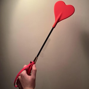May include: A red heart-shaped paddle with a black handle and a red grip. The paddle is held in a hand against a neutral background. The handle has a gold-coloured accent.