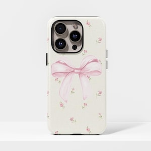 Pode incluir: Capa de telemóvel com fundo creme adornada com pequenas flores cor-de-rosa. Um grande laço rosa em estilo aguarela está centrado na capa. A capa tem uma borda preta e um módulo de câmara.