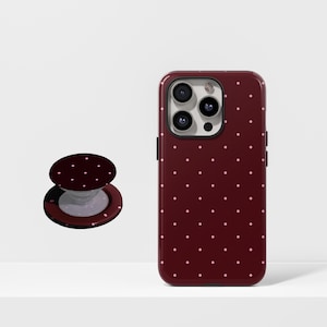 Op de afbeelding: Een bordeauxrode telefoonhoes en bijpassende telefoonhouder met een wit stippenpatroon. De telefoonhoes zit rechts en de telefoonhouder links. De telefoonhoes heeft een camerabult en zijknoppen.