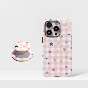 May include: A phone case and matching phone grip featuring a pink gingham pattern with colorful star accents. The phone case is designed to fit a smartphone, and the grip is a round, pop-out style.