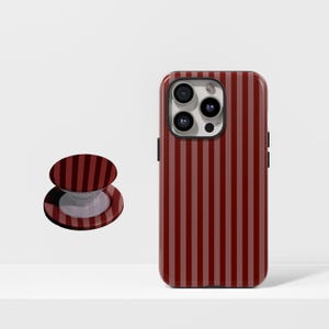 May include: A red and maroon striped phone case and matching phone grip. The phone case is on a white surface. The phone case has a camera and side buttons. The phone grip is round and has a white base.