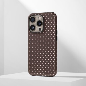 May include: A brown phone case with a repeating pattern of small, evenly spaced pink polka dots. The case has a glossy finish and a camera cutout on the upper left corner. The phone case is designed to fit a smartphone.