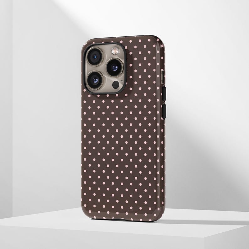 May include: A brown phone case with a repeating pattern of small, evenly spaced pink polka dots. The case has a glossy finish and a camera cutout on the upper left corner. The phone case is designed to fit a smartphone.