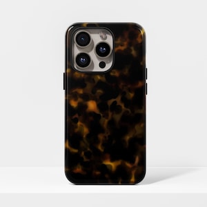 Peut inclure: Une coque de smartphone avec un motif écaille de tortue dans des tons bruns et ambrés. L'étui a une finition brillante et un bord noir. Les lentilles de la caméra sont visibles en haut.