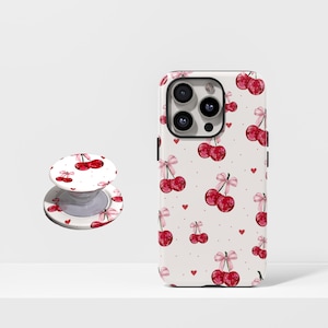 May include: A white phone case and a matching phone grip featuring a pattern of red cherries with pink bows and small red hearts. The phone case is on a white surface.
