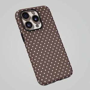 May include: A brown phone case with a pattern of small, evenly spaced pink polka dots. The case has a glossy finish and a camera cutout. The phone case is designed to fit a smartphone.