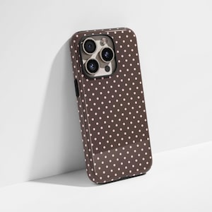 May include: A brown phone case with a glossy finish and a pattern of small, evenly spaced pink polka dots. The case has a camera cutout and black side buttons. The phone case is leaning against a white wall.