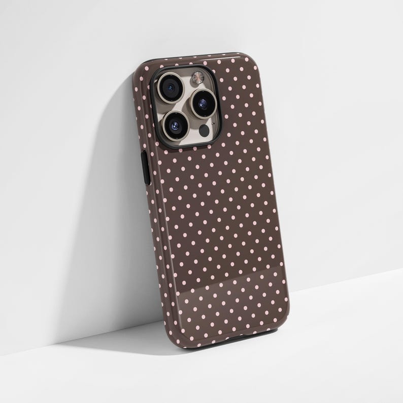 May include: A brown phone case with a glossy finish and a pattern of small, evenly spaced pink polka dots. The case has a camera cutout and black side buttons. The phone case is leaning against a white wall.