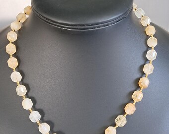 May include: A necklace with faceted, light-colored beads strung on a gold-colored chain. The beads have a mix of peach and clear tones. The necklace is displayed on a black mannequin bust.