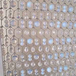 May include: A decorative beaded curtain with a wire mesh backing. The curtain features clear and opalescent beads in various shapes, including round and teardrop, creating a shimmering effect. The beads are strung together, forming a delicate, elegant design.