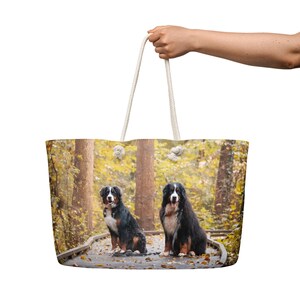 Puede incluir: Un bolso tote grande con una imagen de dos perros de montaña berneses sentados en un camino de madera en un bosque. Los perros tienen pelaje negro, blanco y marrón. El bolso tiene asas de cuerda blanca.