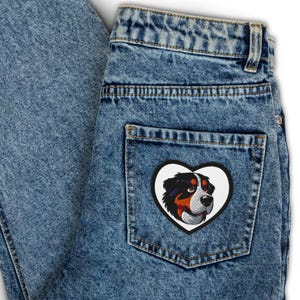 May include: Blue denim jeans with a heart-shaped patch featuring a Bernese Mountain Dog's head. The patch has a white border and showcases the dog's black, brown, and white fur. The jeans are faded and have a classic five-pocket design.
