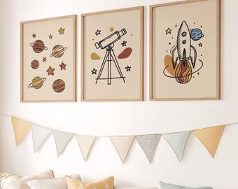 May include: Three framed art prints with space-themed illustrations. One print features planets and stars, another a telescope, and the third a rocket ship. A banner of triangle flags hangs below, with a sofa and stuffed animals in the foreground.