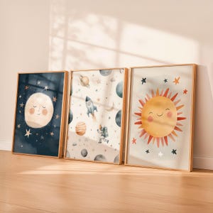 May include: Three framed watercolor art prints featuring celestial themes. The prints depict a smiling moon, a rocket ship with planets, and a smiling sun. The artwork has a light color palette with gold frames.