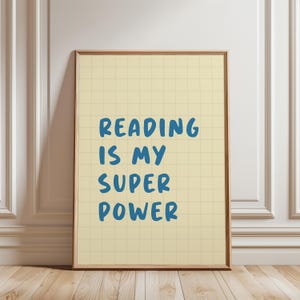 May include: A framed print with the text "READING IS MY SUPER POWER" in blue on a light yellow grid background. The frame is a warm brown colour, and the print is leaning against a white wall.