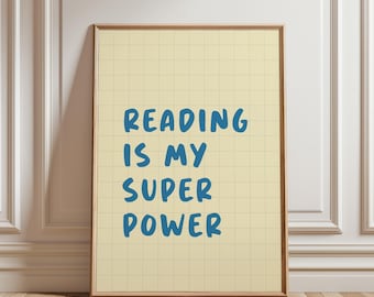 Puede incluir: Un cartel enmarcado con el texto "READING IS MY SUPER POWER" en azul sobre un fondo de cuadrícula amarillo claro. El marco es de color marrón cálido y el cartel está apoyado contra una pared blanca.
