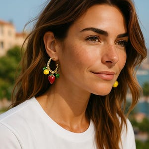 May include: Gold hoop earrings with a fruit charm design. One earring features a red strawberry, a yellow lemon, and green lime charms. The other earring has a single yellow lemon charm. The earrings are worn by a person wearing a white t-shirt.