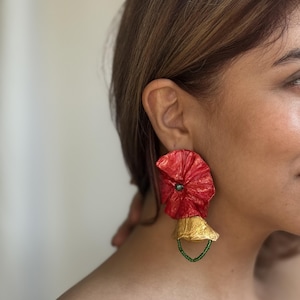 May include: A statement earring featuring a large, crinkled red circular element with a dark center bead, attached to a smaller, textured gold-toned bell-shaped piece. A delicate loop of small green beads hangs from the bottom of the gold component.