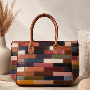May include: A rectangular tote bag constructed from a patchwork of leather strips in various colors including brown, navy, burgundy, olive green, mustard yellow, and cream. It features two brown leather handles attached with brass-toned rivets.