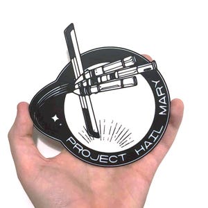 Pode incluir: Um autocolante a preto e branco com o texto "PROJECT MARY HAIL" a rodear um desenho central. O desenho apresenta uma mão estilizada a segurar uma caneta, com um padrão de raios de sol.