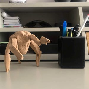 May include: A tan, multi-legged creature figurine with a textured, segmented body and a central head structure, positioned next to a black rectangular pen holder containing several pens and a pencil on a flat surface.
