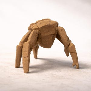 Peut inclure: Une sculpture stylisée de couleur beige représentant une araignée. L'araignée a un corps segmenté et huit pattes, toutes de la même teinte beige. La sculpture est posée sur un fond uni blanc cassé.