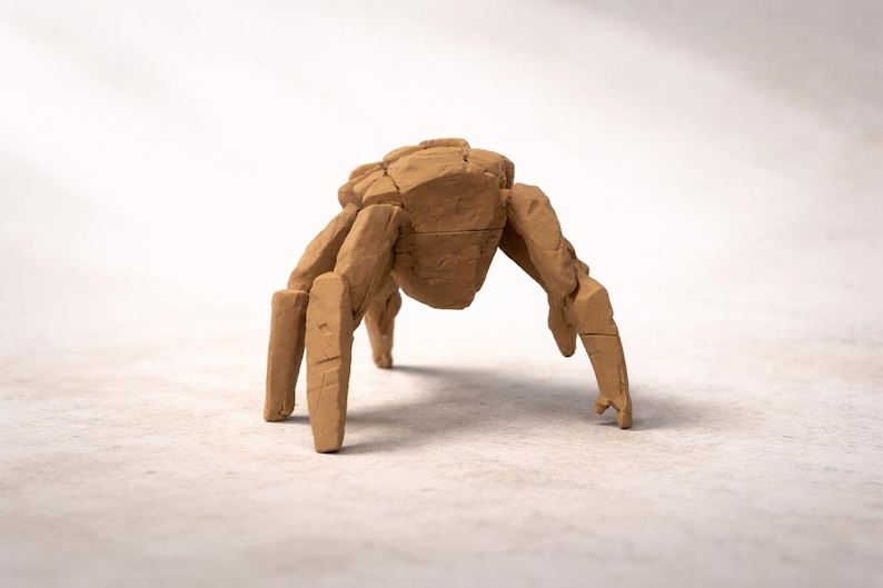 May include: A light brown, clay-like sculpture of a multi-legged creature with a segmented, rock-like body and four visible, thick, angular legs. The surface has a matte, textured appearance with visible cracks and joints between the segments.
