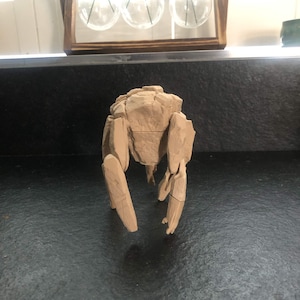 May include: A light brown, multi-jointed mechanical figure with a textured, rock-like exterior. The object features a central torso section with segmented limbs extending downward, displaying visible panel lines and a matte, earth-toned finish.