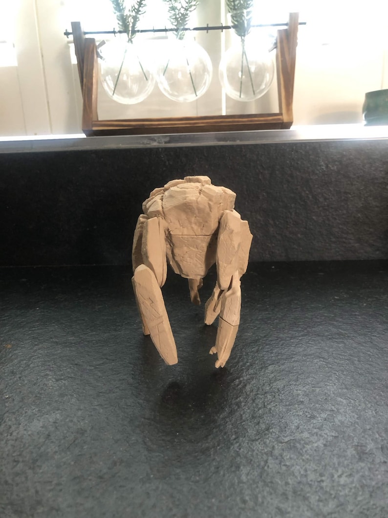 May include: A light brown, multi-jointed mechanical figure with a textured, rock-like exterior. The object features a central torso section with segmented limbs extending downward, displaying visible panel lines and a matte, earth-toned finish.