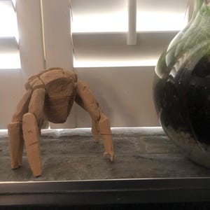 May include: A light tan, multi-faceted crab figurine crafted with geometric, angular planes. The sculpture features a central body section with several articulated, segmented legs extending outward, mimicking the posture of a crustacean.