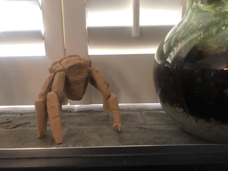 May include: A light tan, multi-faceted crab figurine crafted with geometric, angular planes. The sculpture features a central body section with several articulated, segmented legs extending outward, mimicking the posture of a crustacean.