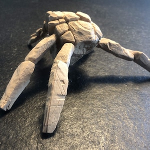 May include: A light beige, multi-legged sculpture with a textured, cracked surface resembling assembled stone fragments. The object features a central cluster of blocky, irregular shapes with several elongated, segmented limbs extending outward.