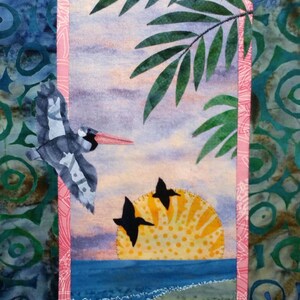 May include: A textile artwork showing a coastal scene with a pelican in flight and two birds silhouetted against a yellow sun. Palm fronds frame the sunset over the ocean. The artwork is framed with a pink border.