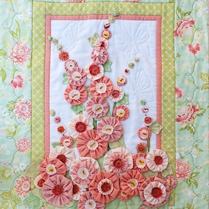 May include: A floral quilt with a central panel of appliquéd fabric flowers in pink and peach hues. The quilt is framed by a green and pink floral border, set against a light green background with a floral pattern. The quilt is handmade.