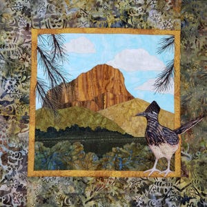 May include: A handmade quilt featuring a landscape scene with a roadrunner. The quilt depicts a mountain, blue sky with clouds, and a roadrunner bird. The quilt has a gold border and a patterned fabric background.