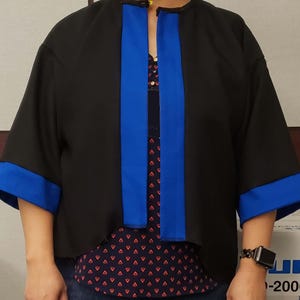 May include: Black jacket with blue trim on the sleeves and front. The jacket has a loose, boxy fit with three-quarter sleeves. The jacket is worn over a patterned top.