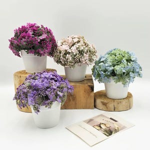 May include: Four small potted plants with colorful faux flowers. The flowers are in shades of purple, pink, and blue. The white pots sit on wooden blocks and a small card is in the foreground.