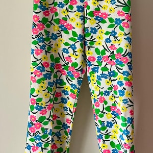 Puede incluir: Pantalones blancos con un vibrante estampado floral. El dise&ntilde;o presenta flores rosas, azules y amarillas con hojas verdes sobre un fondo blanco. Los pantalones est&aacute;n colgados en una percha de madera.