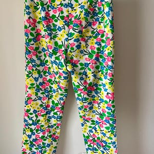 Puede incluir: Pantalones blancos con un estampado floral vibrante con flores rosas, azules y amarillas con hojas verdes. Los pantalones son de corte recto y cuelgan de una percha.
